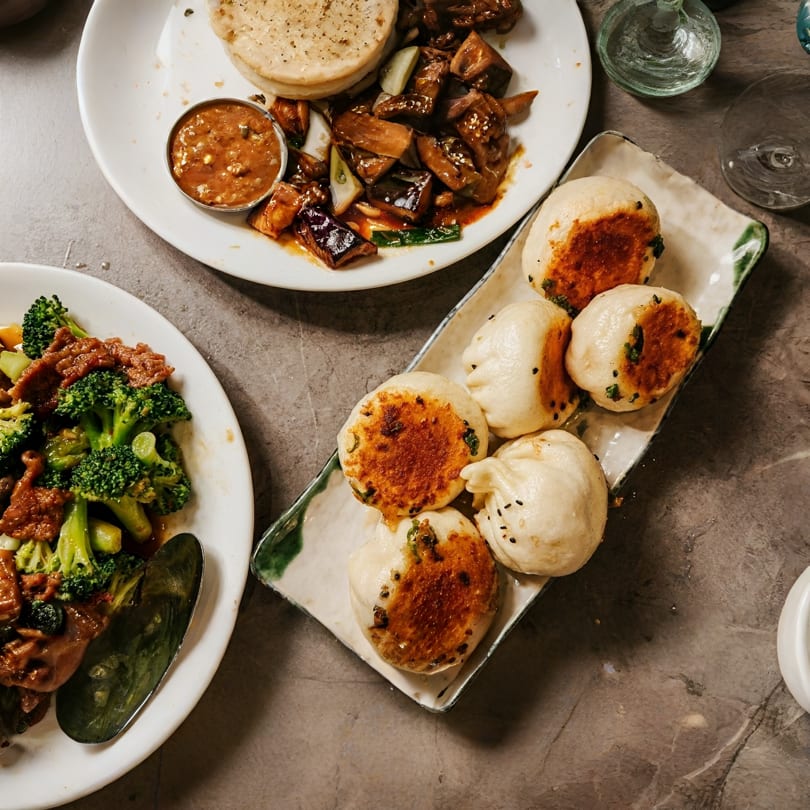 Overhead view of three plates of Asian cuisine, including pan-fried buns, beef and broccoli, and eggplant.