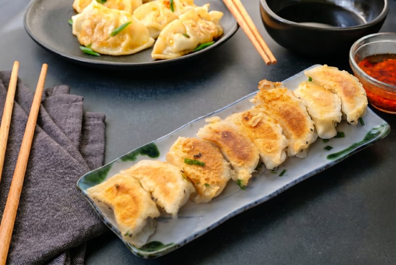 Two plates of golden-brown pan-fried dumplings with dipping sauce and chopsticks on a dark surface.