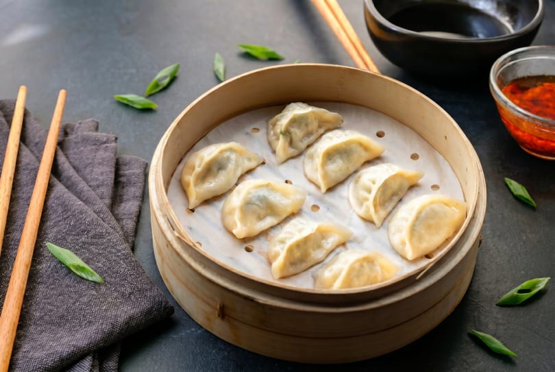 Steamed dumplings in a bamboo steamer with chopsticks, a black bowl, and chili oil.