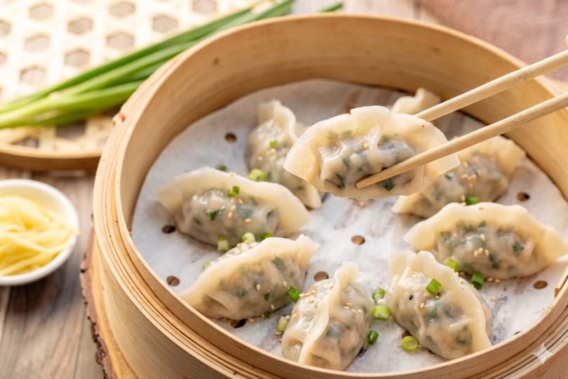 Chopsticks lift a steamed dumpling from a bamboo steamer filled with more dumplings, ginger, and scallions.